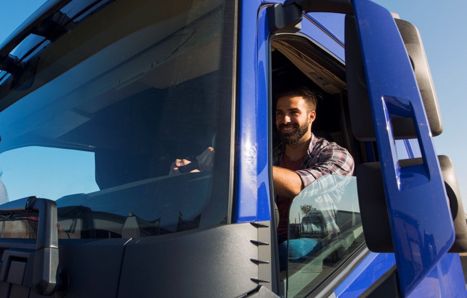 Caminhoneiro sorridente apoia o braço na janela aberta de sua cabine azul, com céu ensolarado ao fundo, transmitindo confiança e satisfação na profissão.