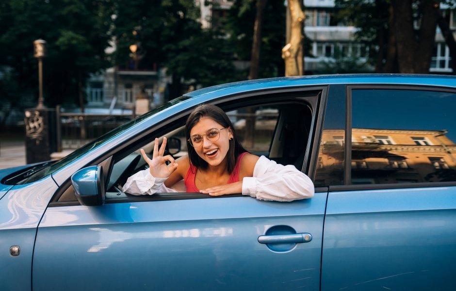 Mulher sorridente dentro de um carro azul, olhando pela janela e fazendo o sinal de 'ok' com a mão.