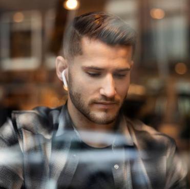 Homem jovem de cabelo curto e barba bem aparada, vestindo camisa xadrez escura. Ele está olhando para baixo com expressão séria e usa um fone de ouvido sem fio branco na orelha direita. A foto foi tirada através do vidro de uma janela, que reflete luzes desfocadas ao fundo.