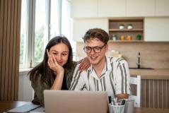 Um casal jovem e sorridente olha atentamente para a tela de um notebook em uma cozinha moderna e iluminada.