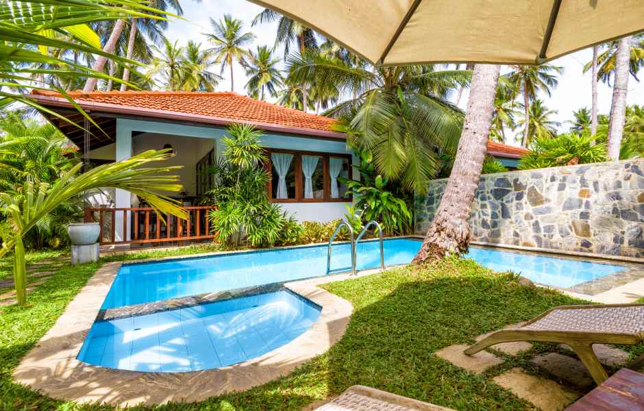 Uma casa de férias com piscina azul, cercada por gramado, coqueiros e uma parede de pedra rústica.