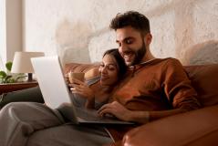 Um casal sorridente relaxa em um sofá de couro enquanto usa um notebook e toma café em um ambiente iluminado.