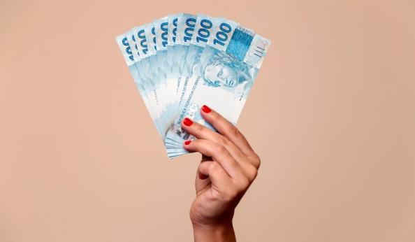Mão segurando várias cédulas de dinheiro de 100 reais, destaque para a cor azul das notas e a expressão de valor e riqueza.