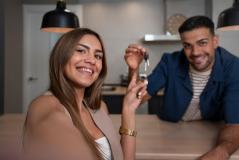 Um casal jovem e sorridente comemora a conquista da casa nova enquanto a mulher tira uma selfie. O homem segura as chaves em destaque ao fundo, em uma cozinha moderna com iluminação aconchegante.
