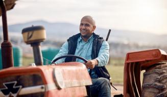Agricultor sorridente conduz um trator vermelho no campo, com paisagem rural e montanhas ao fundo, transmitindo satisfação e orgulho no trabalho no campo.