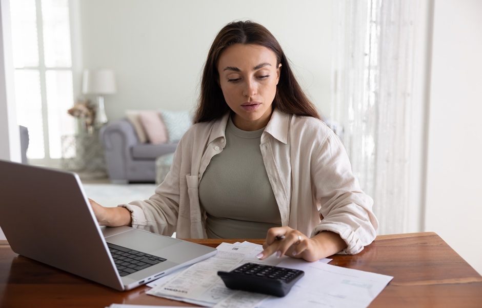 Mulher estudando contas financeiras em casa, com laptop, calculadora e papéis sobre a mesa, em um ambiente doméstico bem iluminado, ideal para gerenciamento financeiro.
