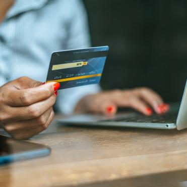 Close de mulher segurando cartão de crédito azul enquanto digita no laptop, realizando um pagamento online com celular ao lado sobre a mesa.