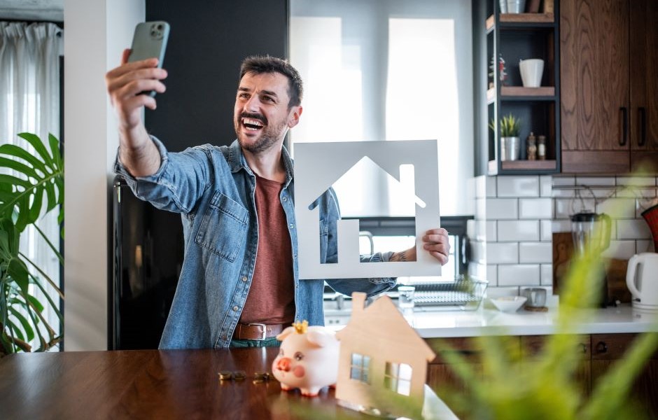 Homem feliz tirando selfie na cozinha moderna enquanto segura uma casa de papelão com uma decoração aconchegante e plantas ao redor.