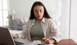 Mulher estudando contas financeiras em casa, com laptop, calculadora e papéis sobre a mesa, em um ambiente doméstico bem iluminado, ideal para gerenciamento financeiro.